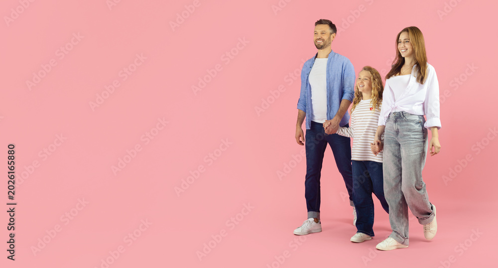 Naklejka premium Happy caucasian family of three walking together on pink studio background with copy space. Father, mother and daughter stroll hand in hand in casual outfits looking away