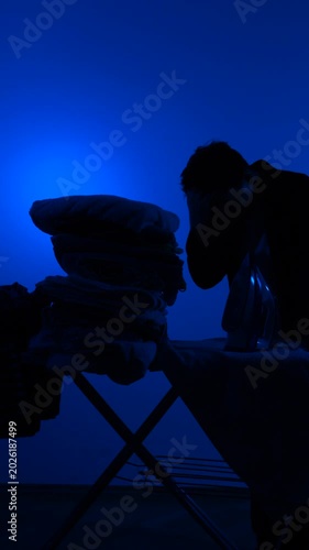 Vertical video man silhouette ironing clothes at board against deep blue backdrop, portraying fatigue and daily household drudgery