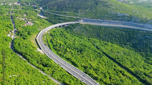 An aerial perspective of a modern highway A7 and viaduct curving through lush green, forested hills in Croatia, bathed in late afternoon sunlight. Hreljin