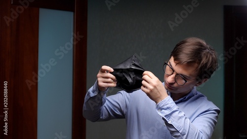 Foto Young man in glasses and a light blue shirt shows an empty wallet, expressing fi