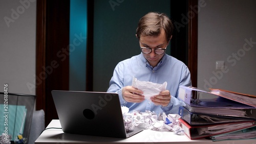 Bild auf Leinwand Young man crumpling paper at his desk, working late in a dimly lit office, exper