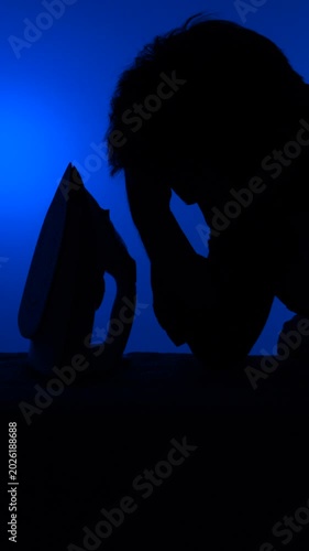 Vertical video man silhouetted ironing with visible fatigue and reluctance against a vibrant blue backdrop, conveying household drudgery