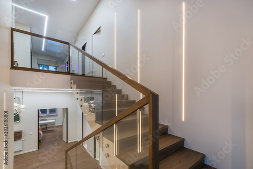 Stairs to the second floor in the lobby of a luxurious mansion. White wall decor, parquet, dark wood staircase with glass railings.