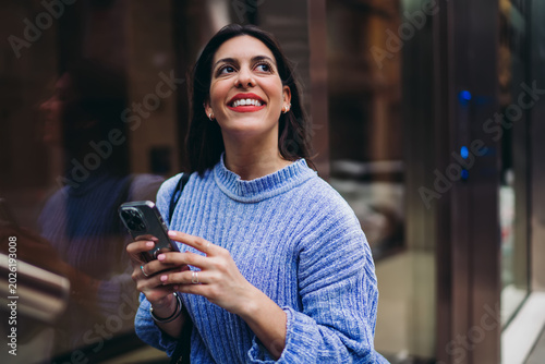 Millennial woman smiling while looking at smartphone screen, receiving joyful message, mobile banking success, digital lifestyle in city, wireless connection, authentic representation