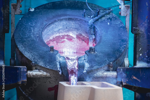 Molten aluminum glowing red as it pours from a large industrial melting furnace into a mold inside a foundry.