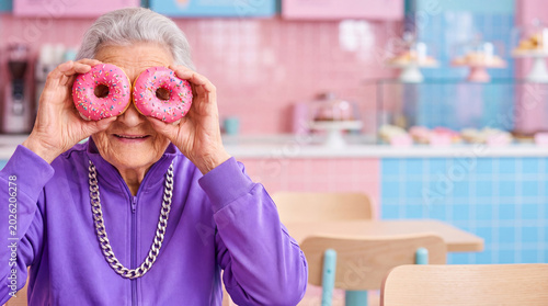 Senior woman playing with pink donuts like glasses in a bakery. Cheerful elderly lady having fun acting like a teenager. Concept of active old age and playful retirement lifestyle.