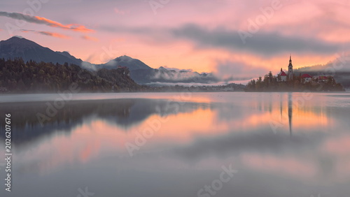 Foto Reddish, foggy sunrise over the town and the Assumption of Mary church on Bled island seen from Velika Zaka pier across Lake Bled