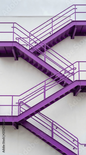 Purple industrial fire escape staircase attached to white building wall. Geometric architectural construction for city safety. Exterior metal stairs as modern urban background element.