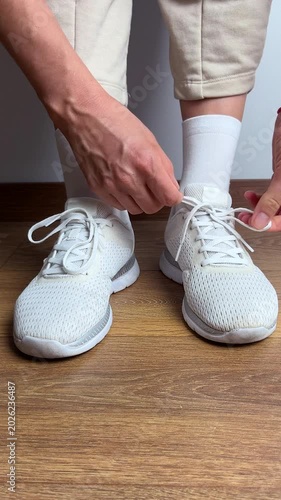 Close up of a woman tying shoelaces on white sneakers indoors. Sport shoes on wooden floor. Preparing for workout, fitness, jogging, training or daily casual activity.