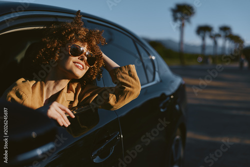 Canvas Print A young woman leans out of a car window, wearing a brown jacket and sunglasses, basking in warm sunlight while palm trees blur by in the background