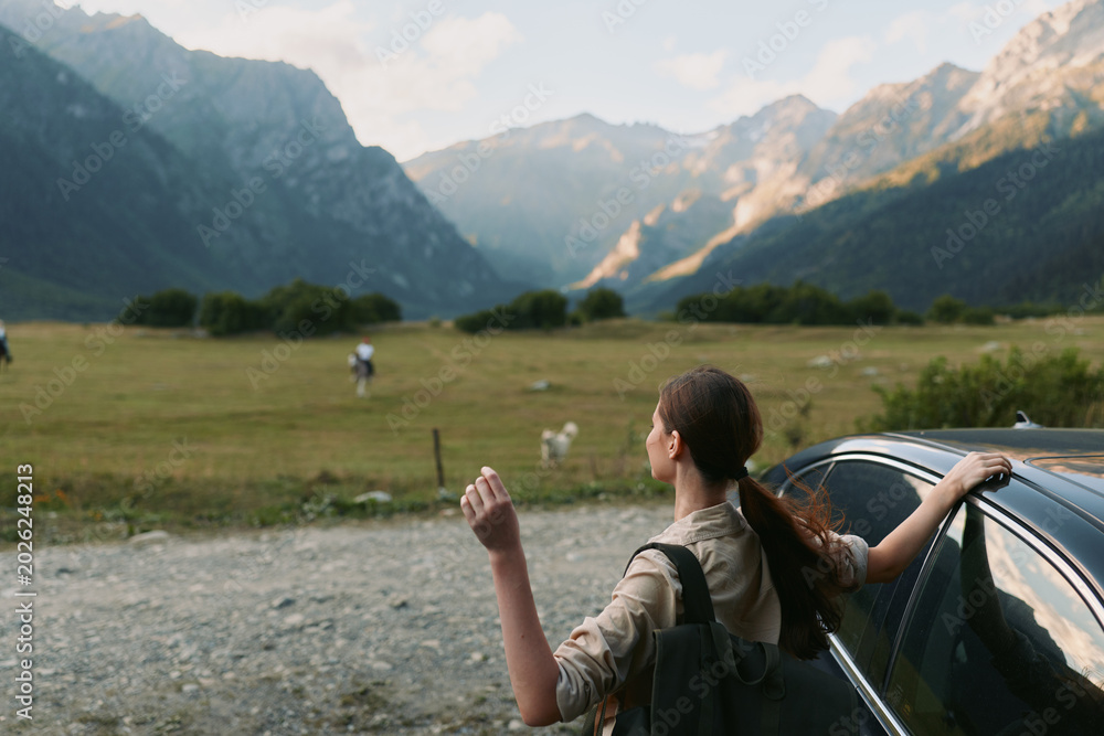 Fototapeta premium Woman traveler by a car in a mountain valley, gazing toward distant peaks, backpack ready, sunlight and open air, capturing a peaceful outdoor lifestyle moment