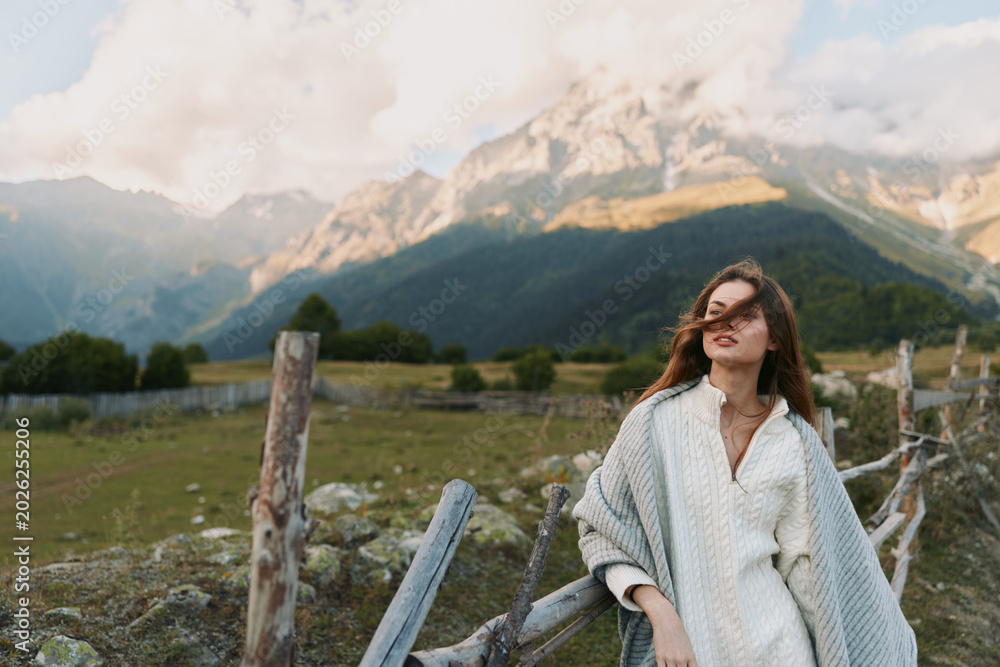 Fototapeta premium Cheerful woman outdoors wearing cozy cardigan near a rustic fence, mountain backdrop, soft sunlight, relaxed lifestyle moment, natural tones, lush landscape
