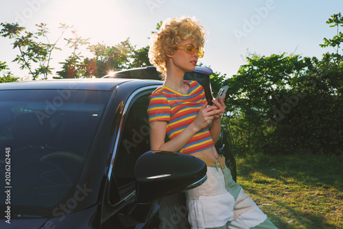 A woman leans against a dark car in a sunny outdoor setting  wearing a striped tee and sunglasses. A retro color palette enhances a casual lifestyle moment.