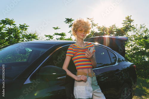 Smiling woman beside a car wearing a striped crop top in bright sunlight  outdoor scene conveying a casual lifestyle  summer vibe  and vintage film warmth  under soft natural lighting.