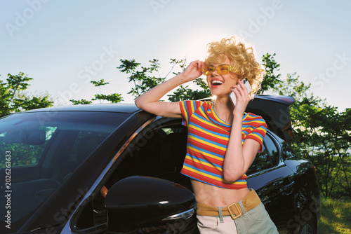 Young woman with curly blonde hair in a striped crop top talks on a mobile phone beside a dark car in a sunny outdoor setting  exhibiting a cheerful mood and casual vibe