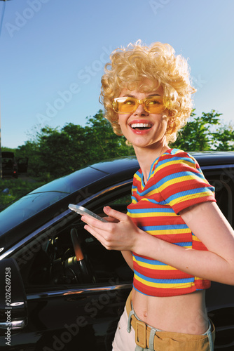 A cheerful woman with retro blond curls and yellow sunglasses stands beside a sleek car in bright sunlight. Colorful striped top highlights a playful  carefree lifestyle.