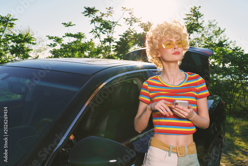 Woman outdoors  retro fashion  striped crop top  standing beside a car in bright sunlight  casual lifestyle  vintage colors  lush green background  carefree moment