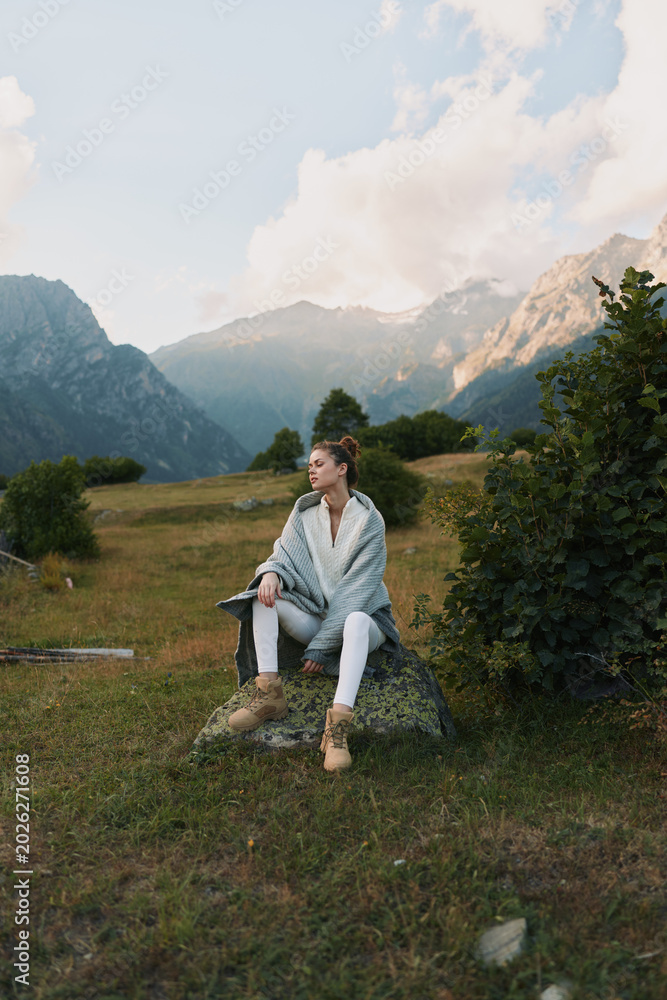 Fototapeta premium A woman sits on a rock in a mountain meadow, wrapped in a soft poncho, enjoying a quiet moment in nature. A serene lifestyle scene of relaxation and exploration.