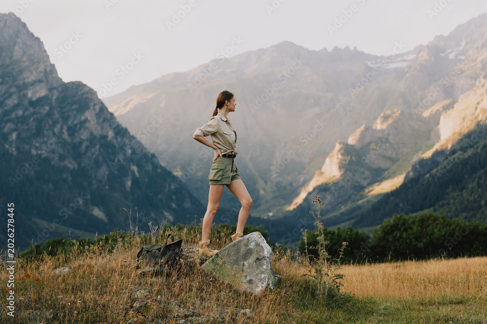 Fototapeta premium Young woman in hiking shorts and beige top stands on a rock amid a rugged mountain valley, enjoying a moment of exploration and freedom with a sense of adventure and calm as the wind sweeps the grass.