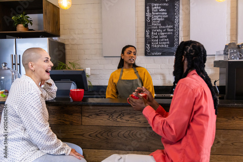 Diverse patrons talking with Indian barista in mustard sweater olive apron at counter with red mugs