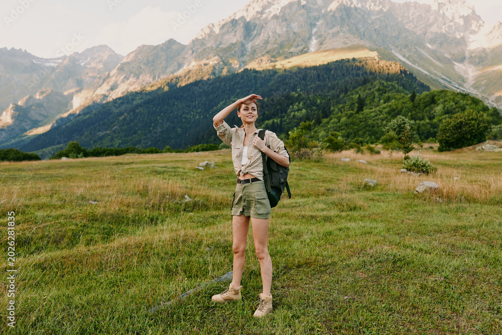 Fototapeta premium Woman with backpack standing in a grassy meadow near mountains, ready for adventure. Outdoor lifestyle scene with natural light and expansive landscape.
