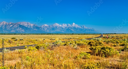 A wide view of the Grand Teton mountain range rising above the dry grassy plains of the Jackson Hole valley, as seen from a quiet highway under a vivid blue summer sky. Wyoming, United States
