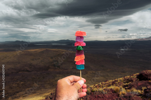 Dramatic storm clouds over a remote Andean valley with a hand holding a vibrant rainbow candy stick.
