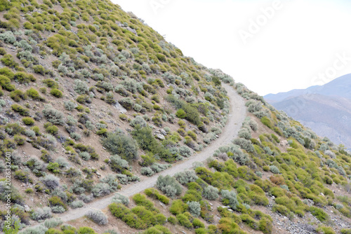 Winding dirt trail on the side of a mountain, another mountain in the background with bright cloudy sky behind