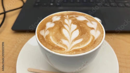 A beautifully decorated cup of fresh cappuccino with intricate latte art, set against a blurred laptop screen in a cozy coffee shop environment.