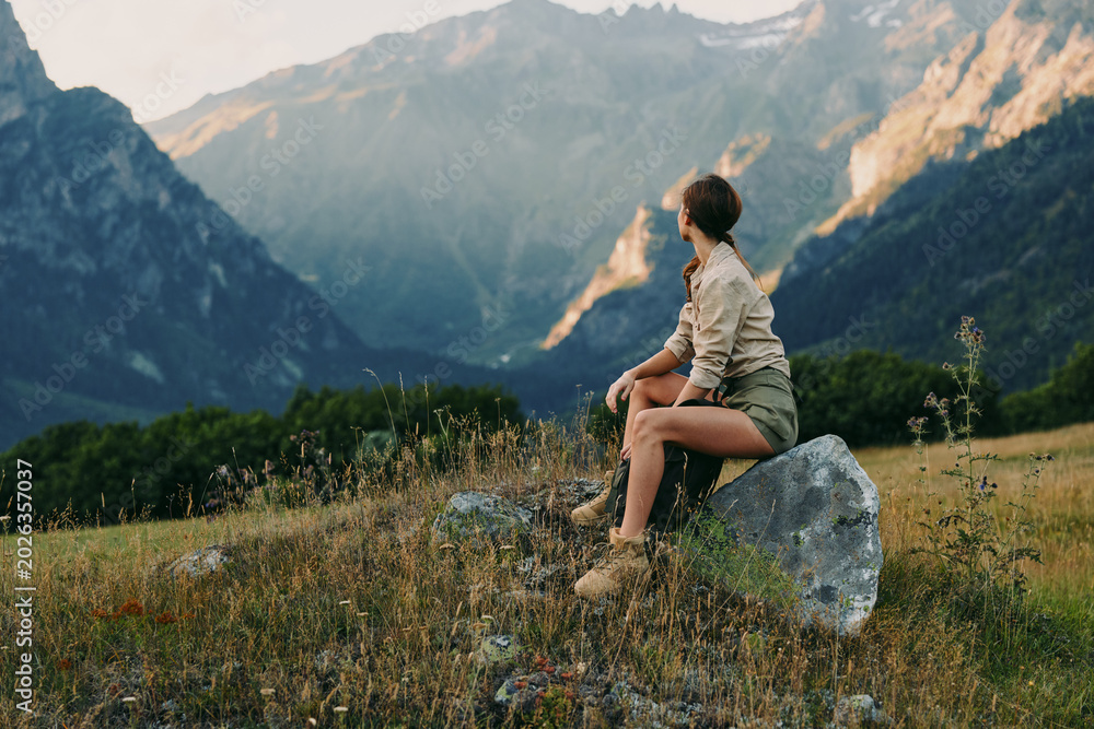 Fototapeta premium Bright outdoor lifestyle scene with a woman sitting on a rock in a meadow, mountains in the background, relaxed pose, natural light, gentle breeze, warm tones