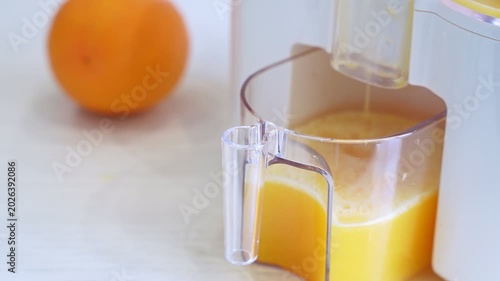 Close-up of fresh orange juice pouring from a white electric juicer into a clear container. A whole orange sits in the background of a bright modern kitchen during the day.