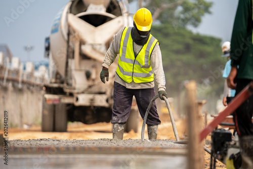 Картината върху платно Road Construction Work: A construction worker in a yellow hard hat and safety vest pours cement on a road construction site, with a concrete mixer truck in the background