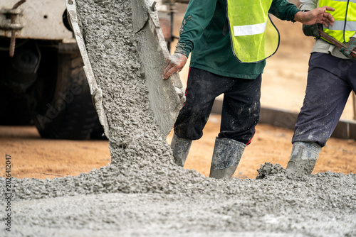 Bild auf Leinwand Pouring Concrete at Construction Site: Two workers in high-visibility vests and boots carefully guide a flow of wet concrete being poured from a truck