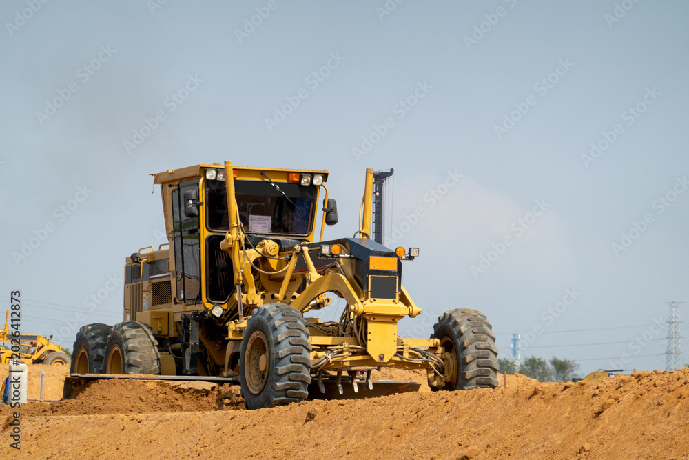 Fototapeta premium Road Grader on Earth: A heavy-duty yellow road grader with large tires levels a mound of dirt at a construction site under a clear sky, showcasing industrial progress.