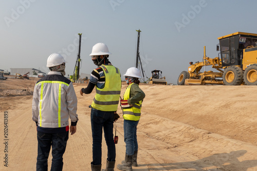 Photos Construction Site Management: Three engineers in hard hats and safety vests oversee operations on a bustling construction site, with heavy machinery like a grader and drilling rigs in the background
