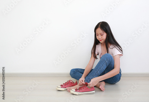 Portrait of young Asian girl with long dark hair in pink t-shirt and blue jeans, sitting on the floor while putting on her red sneakers against clean white wall.