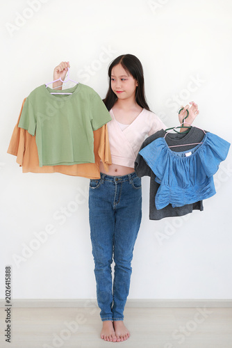Beautiful young Asian girl standing against white wall, holding up various colorful shirts on hangers while deciding what to wear.