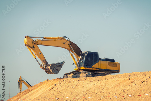Photos Heavy Machinery at Work: A powerful yellow excavator is actively digging into a mound of earth under a clear blue sky, signifying ongoing construction and development