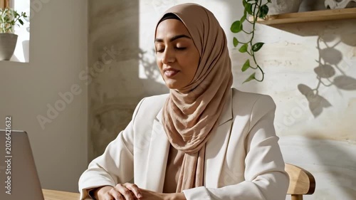 Remote worker home office portrait of confident woman in beige hijab and cream blazer at laptop, sunlit plant shelf workspace, calm modern freelance meeting atmosphere with elegant professional focus