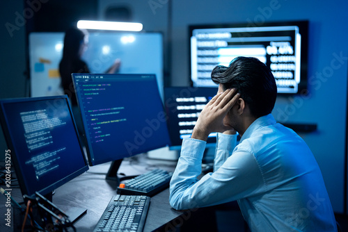 Photos A Stressed programmer has headache while writing program code on multiple desktop screens while his colleague working in the background in a modern office