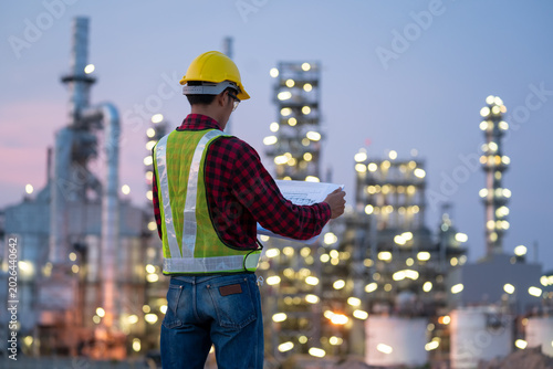 Industrial Overseer at Dusk: An engineer in a yellow hard hat and high-visibility vest reviews blueprints against the backdrop of a sprawling oil refinery at twilight.