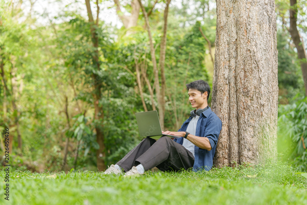 Obraz premium Asian man working remotely on laptop while sitting against tree in outdoor park