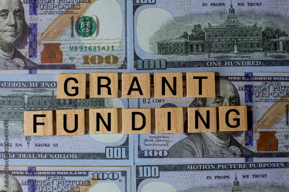 Fototapeta premium Grant Funding Concept with Wooden Letter Tiles on Dollar Bills Background Representing Finance Investment Support