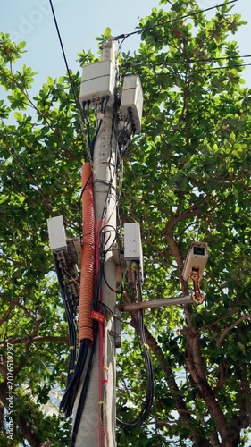Monitoring Equipment On City Utility Poles. Security Camera Installed Amid Trees On Tall Utility Pole. City Street Surveillance Device With Visible Wiring And Natural Green Surroundings