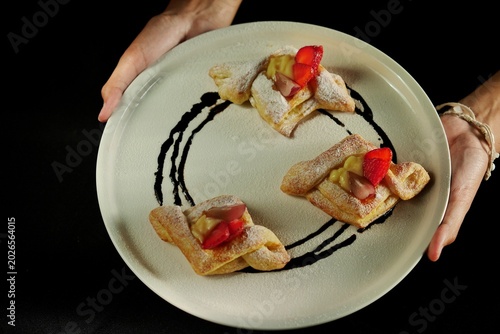 Top view of a plated dessert featuring three pieces of puff pastry topped with custard cream and fresh strawberries, dusted with powdered sugar and decorated with chocolate sauce. 