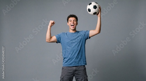 Joyful young man celebrating victory while holding a soccer ball against gray backdrop