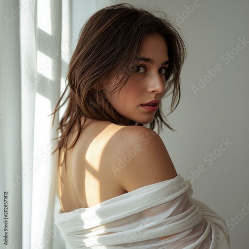 A beautiful young woman with wavy brown bob hair looks back over her bare shoulder while wearing a draped white fabric near a sunlit window with sheer curtains.