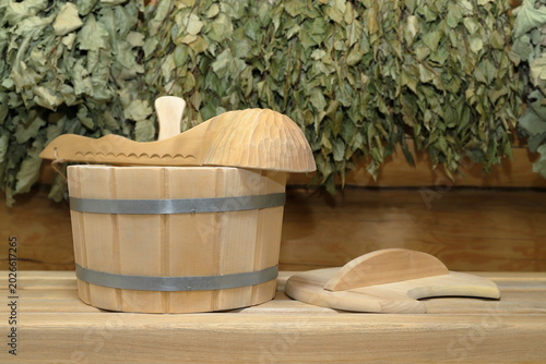 Foto A wooden vat and a wooden ladle are on a plank bench on the background of a log wall with hanging on it a set of a dry birch and oak brooms