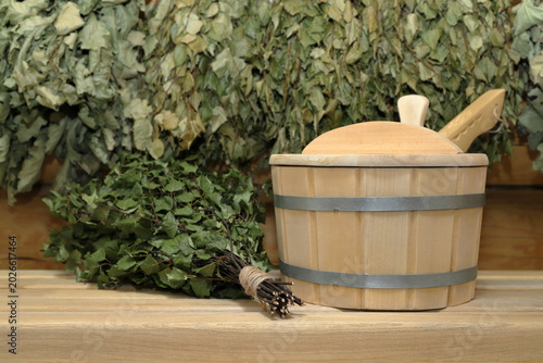 Foto A wooden vat with a wooden ladle and a birch broom are on a plank bench on the background of a log wall with hanging on it a set of a dry birch and oak brooms
