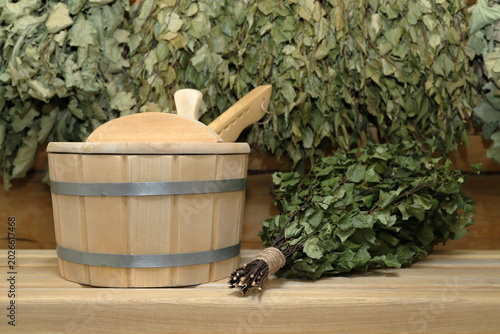 Bild auf Leinwand A wooden vat with a wooden ladle and a birch broom are on a plank bench on the background of a log wall with hanging on it a set of a dry birch and oak brooms